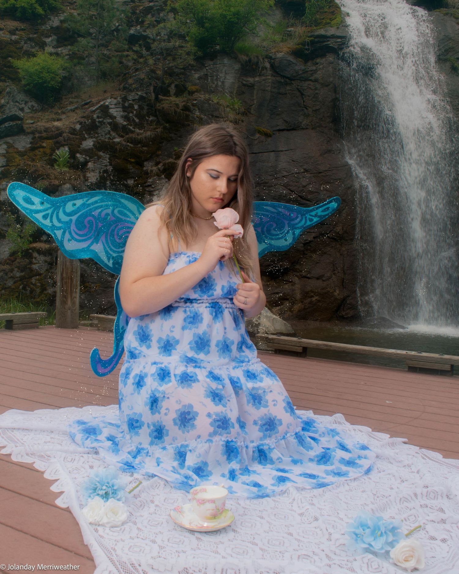 Woman in blue floral dress with butterfly wings smells a rose on a deck near a waterfall.