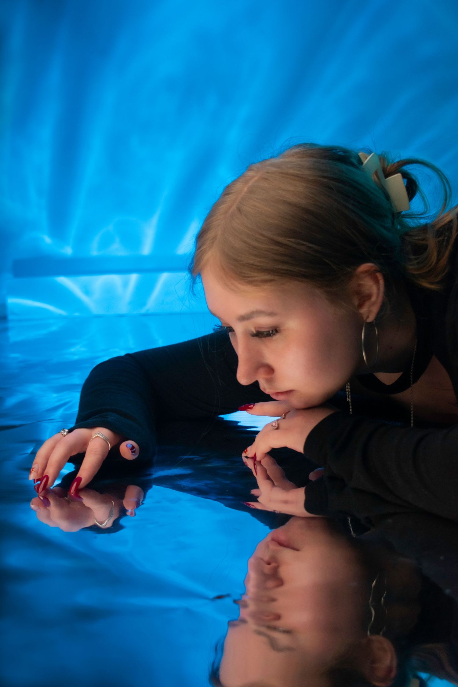 Young woman in black top lying on reflective surface with blue background, looking down with hoop earrings.