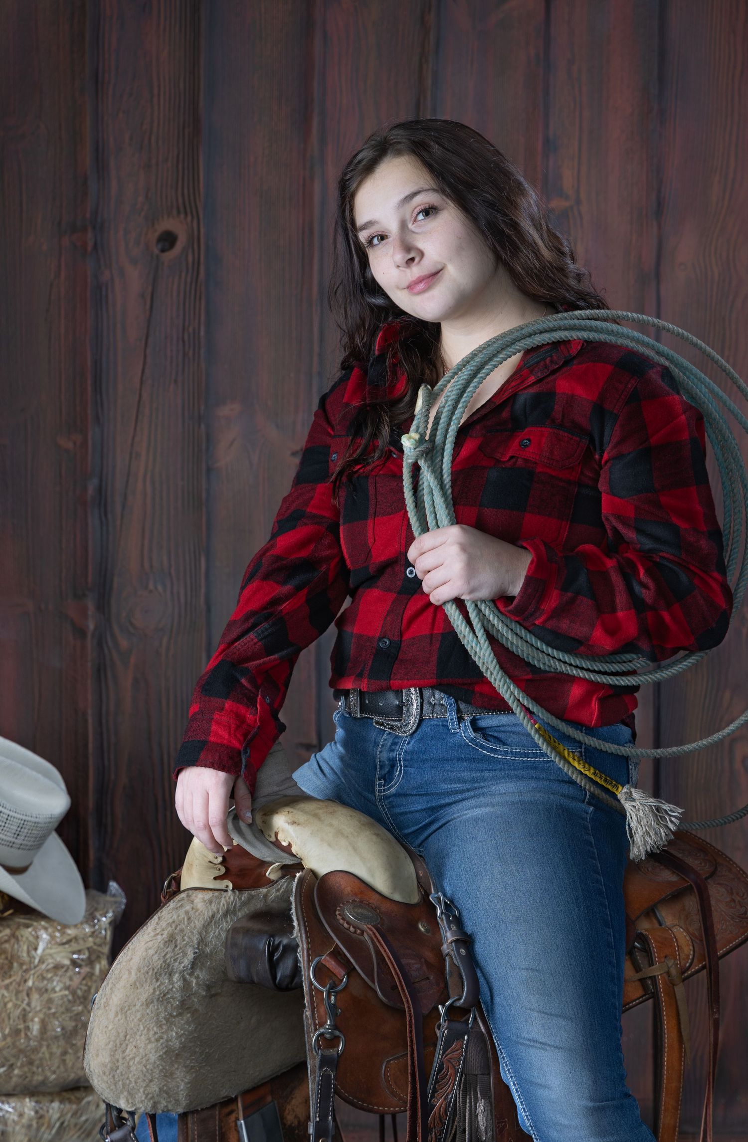 Young woman in red plaid shirt holds a lasso next to a saddle in a rustic wooden setting.