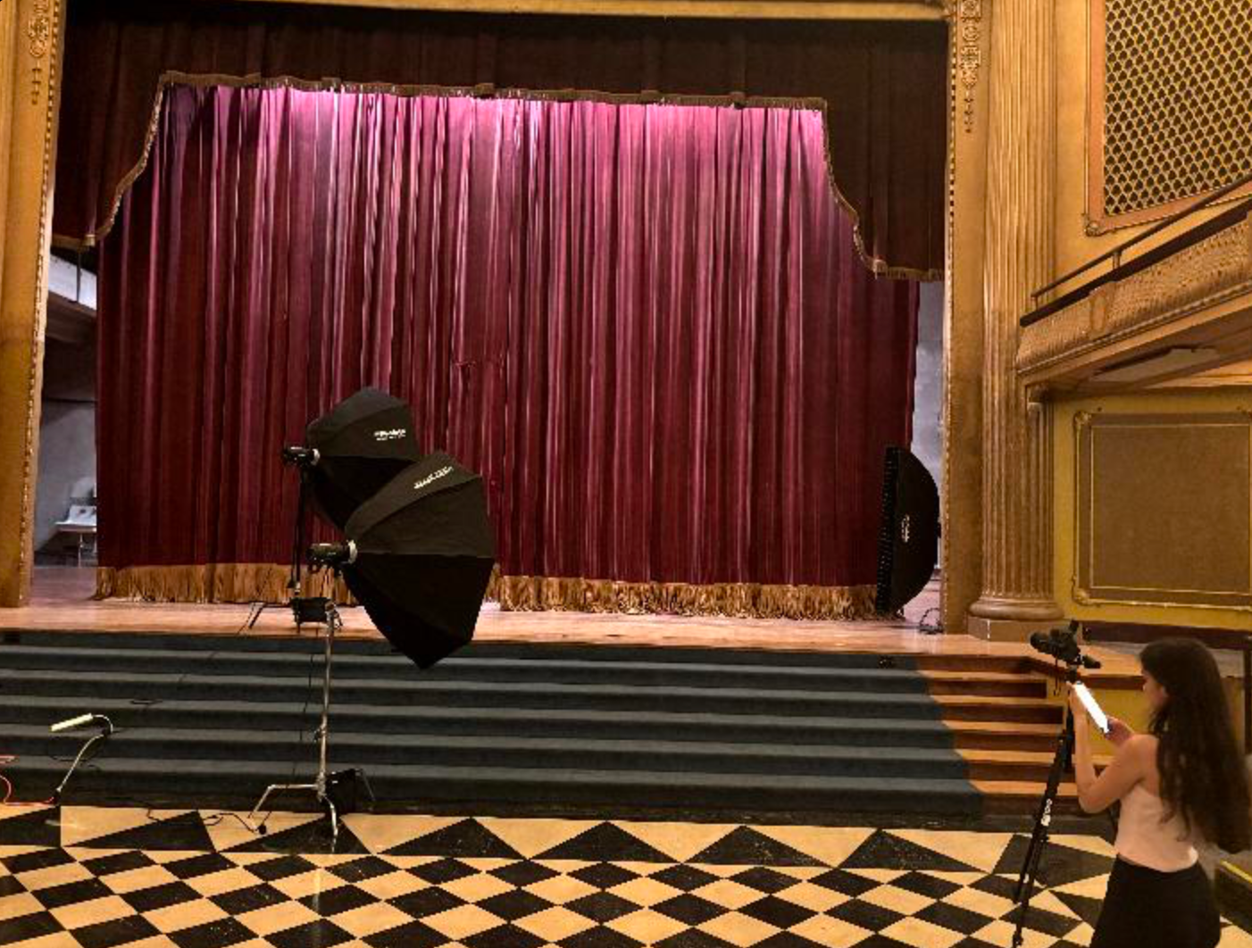Theater stage with red velvet curtains, photography lighting equipment, and checkered floor in an elegant venue.