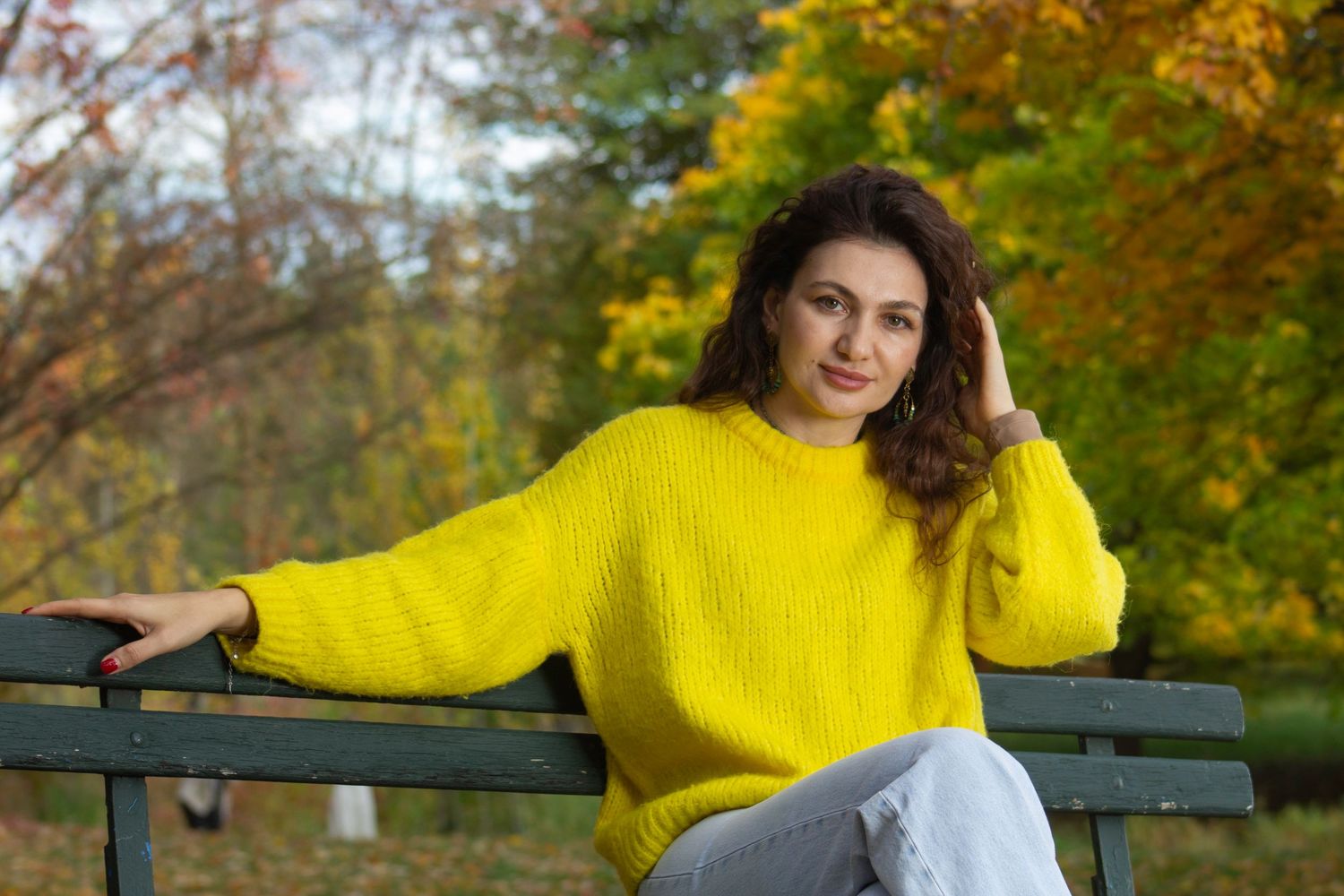Woman in bright yellow sweater sits relaxed on a park bench surrounded by colorful autumn foliage.