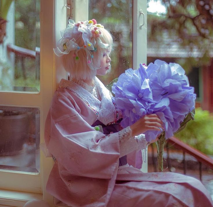 Woman in pink vintage dress holding large lavender paper flower while seated by sunny window.