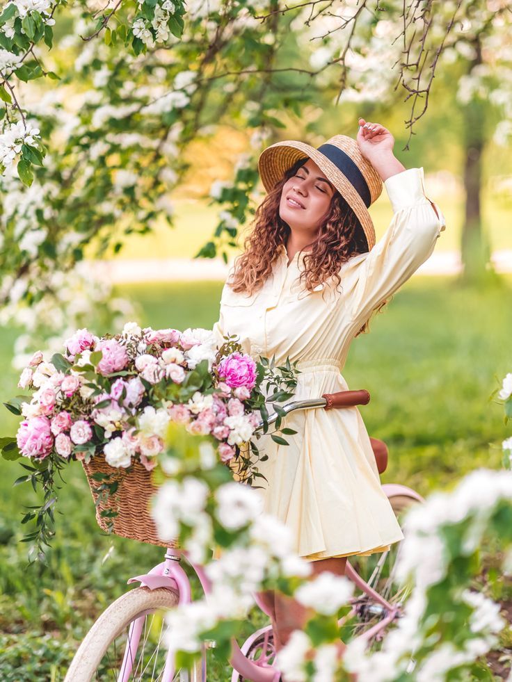 Young woman in yellow dress and straw hat posing with pink bicycle and flower basket in blooming spring garden.