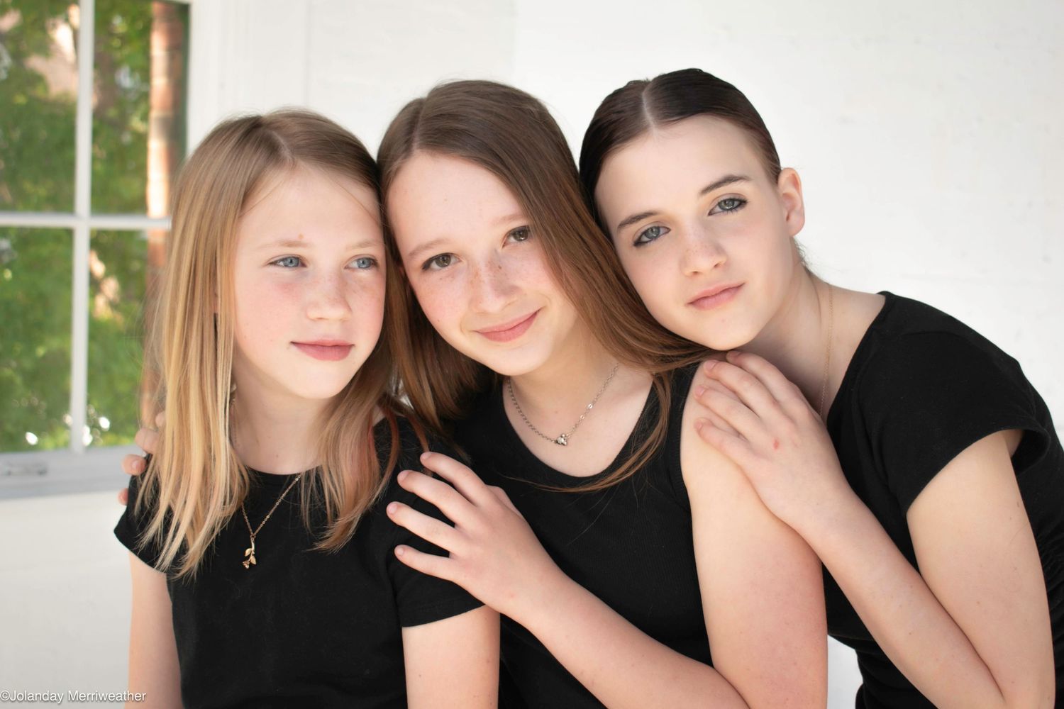 Three young girls in black tops pose together smiling in a bright studio portrait photo.