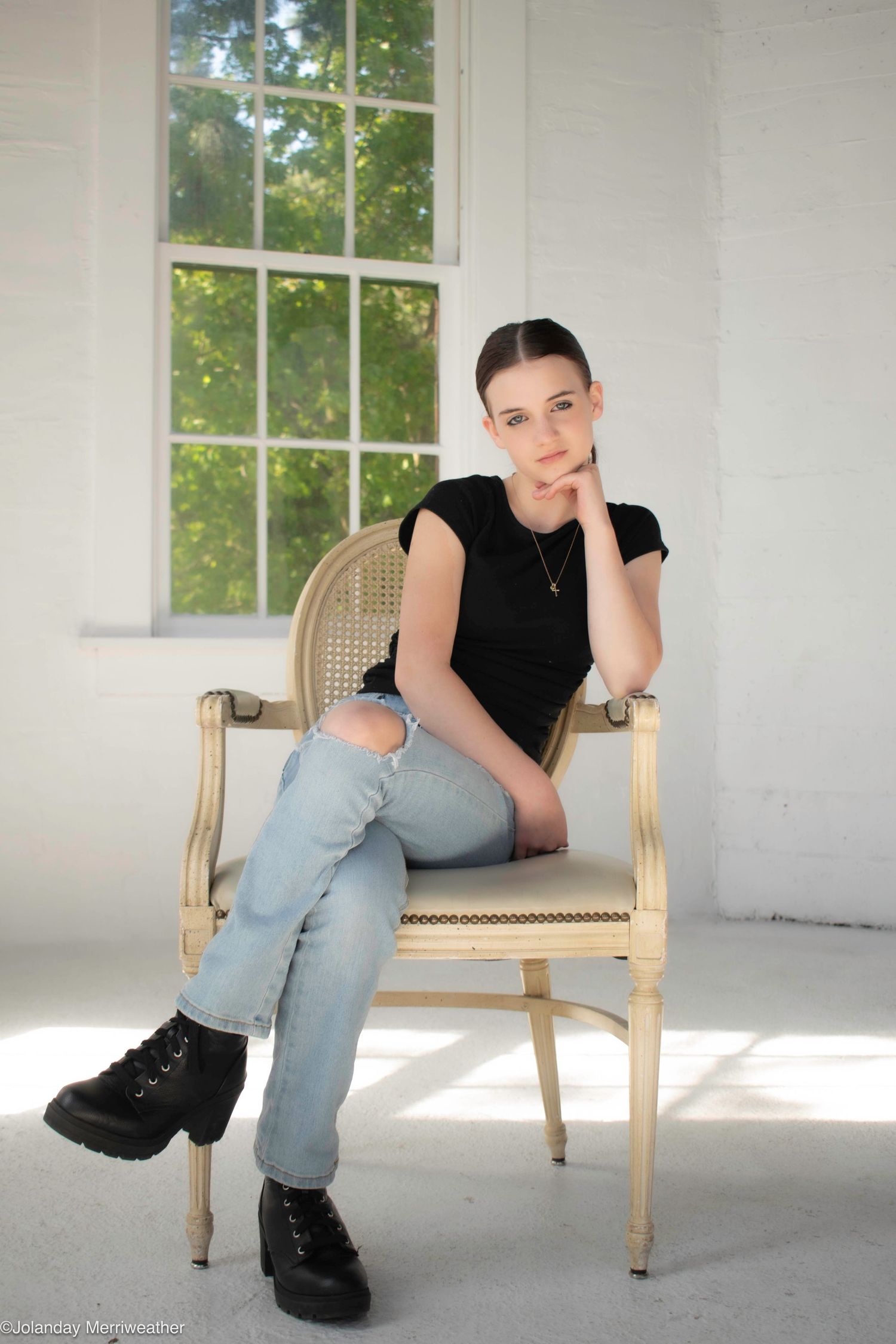Young woman in black top and ripped jeans sits in ornate chair by bright window.