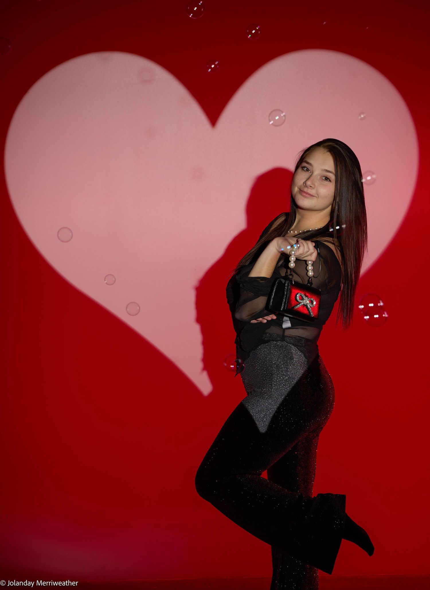 Young woman posing in front of a red heart backdrop, wearing a black outfit, with soap bubbles floating around her.