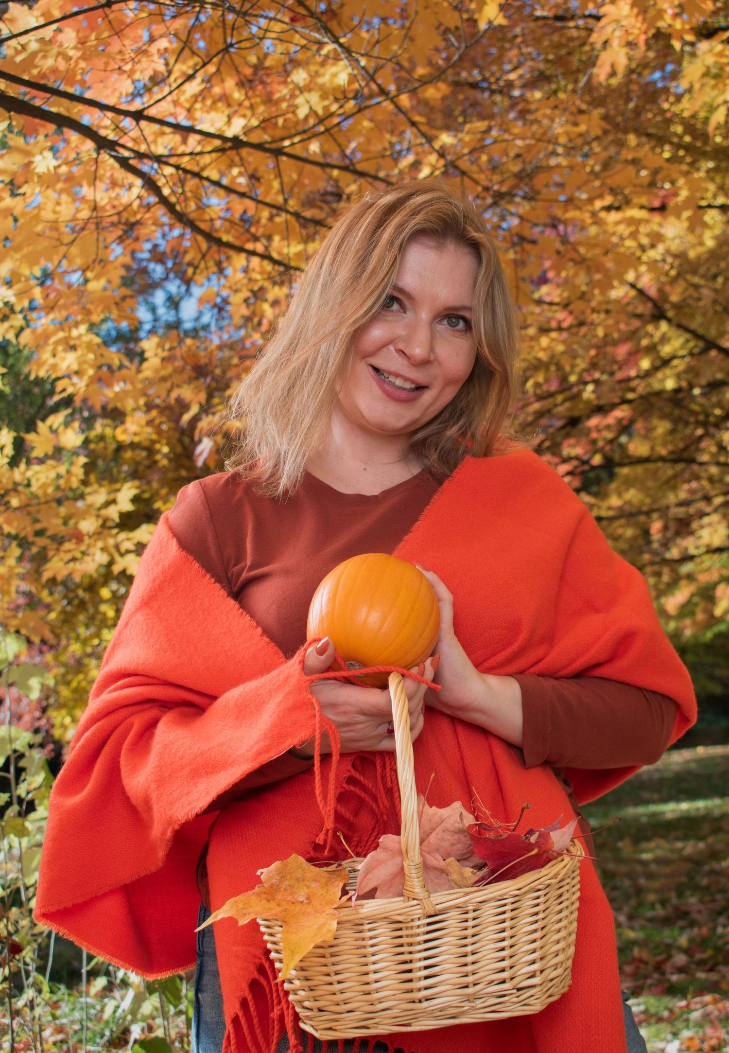 Smiling blonde woman in orange wrap holds a pumpkin and basket of autumn leaves outdoors.