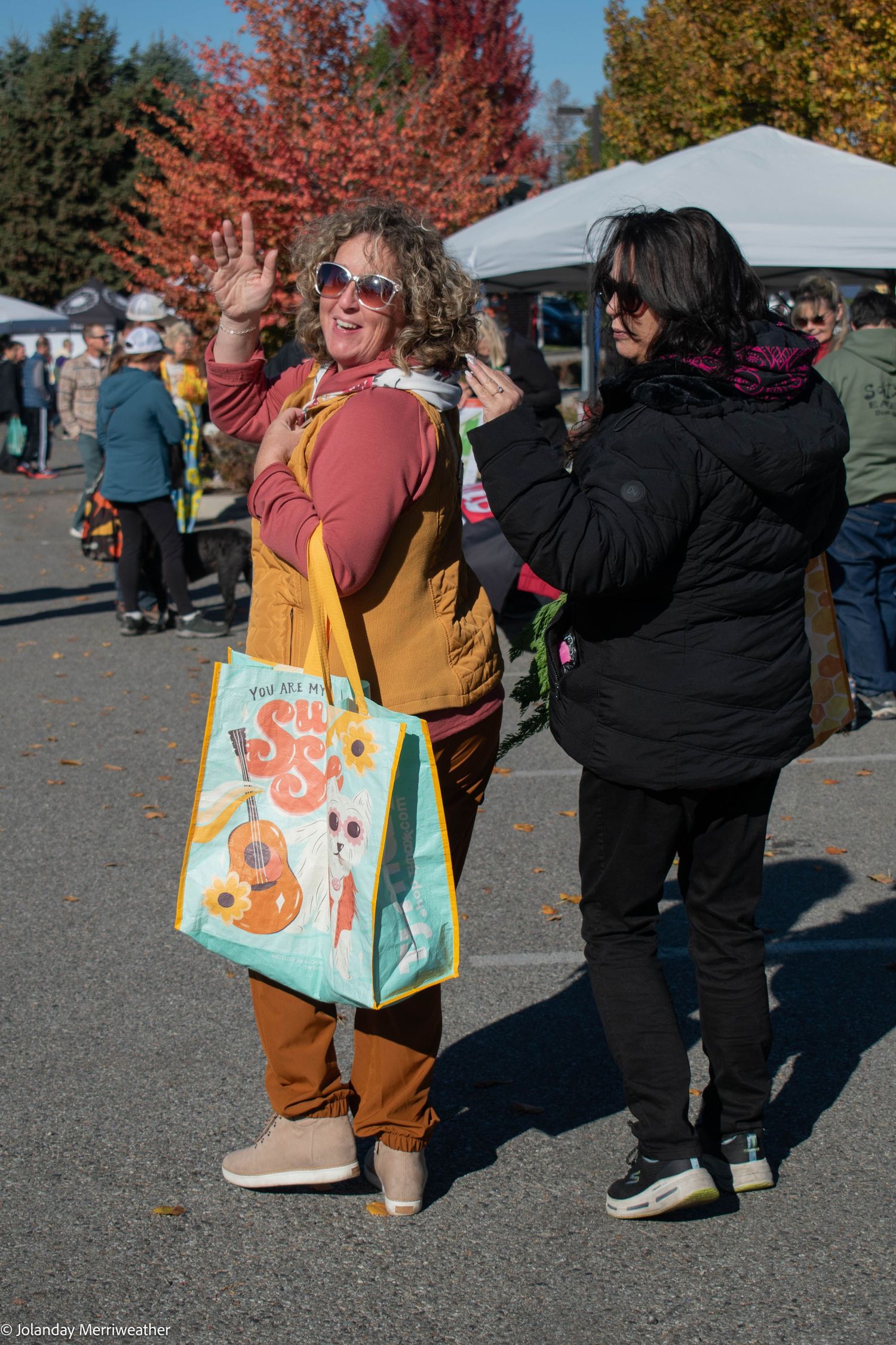 Two women socializing at an outdoor autumn market, one waving and carrying a tote bag.