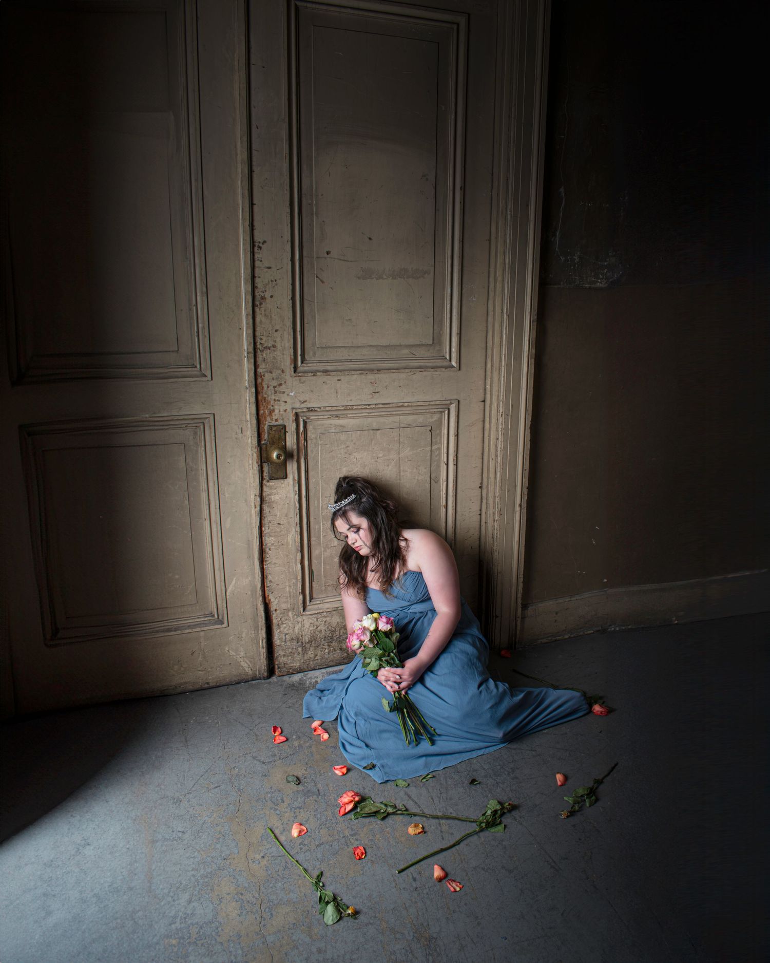 A woman in a blue gown sits on the floor holding roses near a vintage door with scattered petals around her.