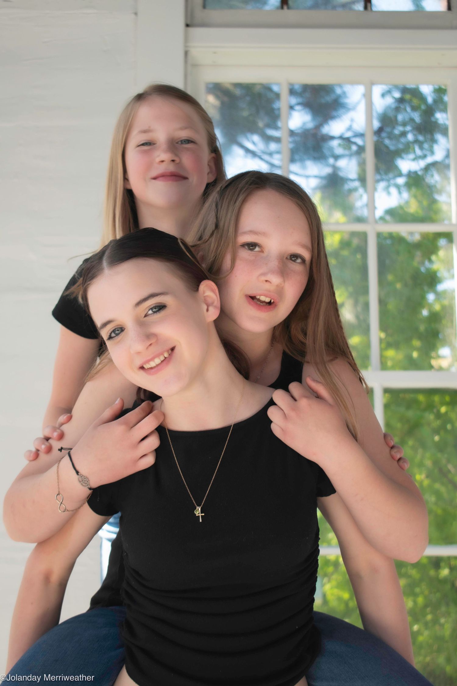 Teen girl in black shirt poses with two younger sisters near sunlit window.