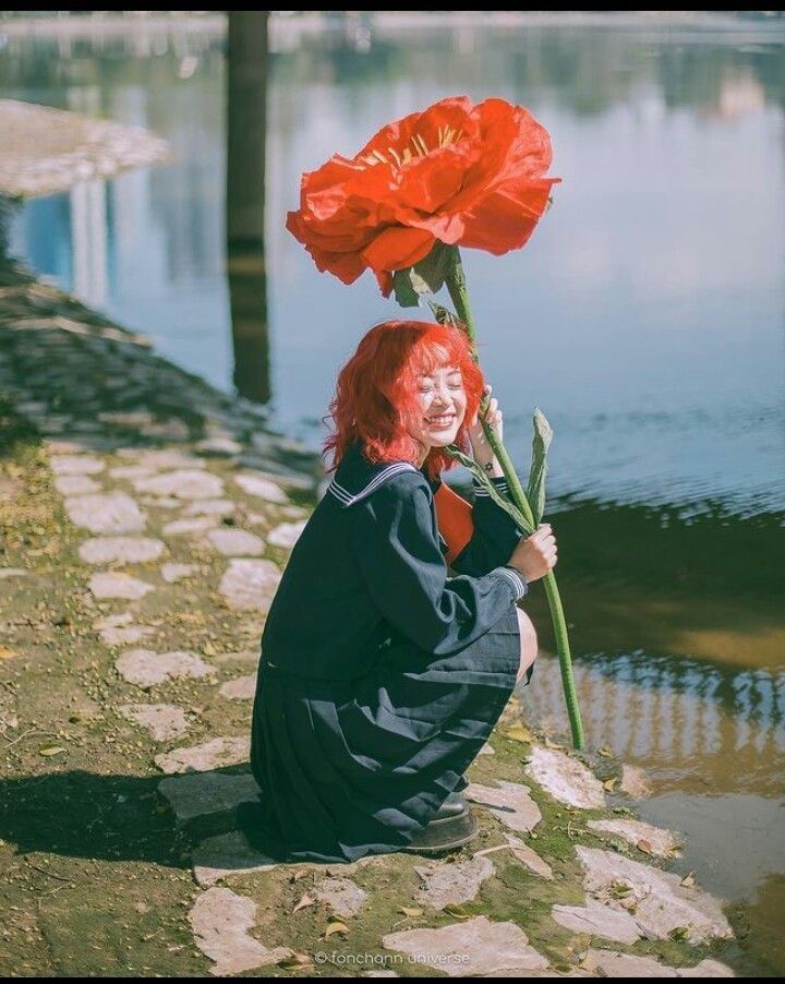 A smiling girl with red hair holds a giant red poppy flower while crouching by a riverside.