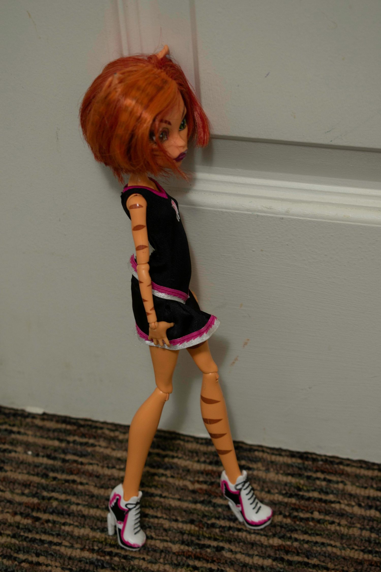 Monster High doll with red hair wearing black dress and sneakers posed against a gray wall.