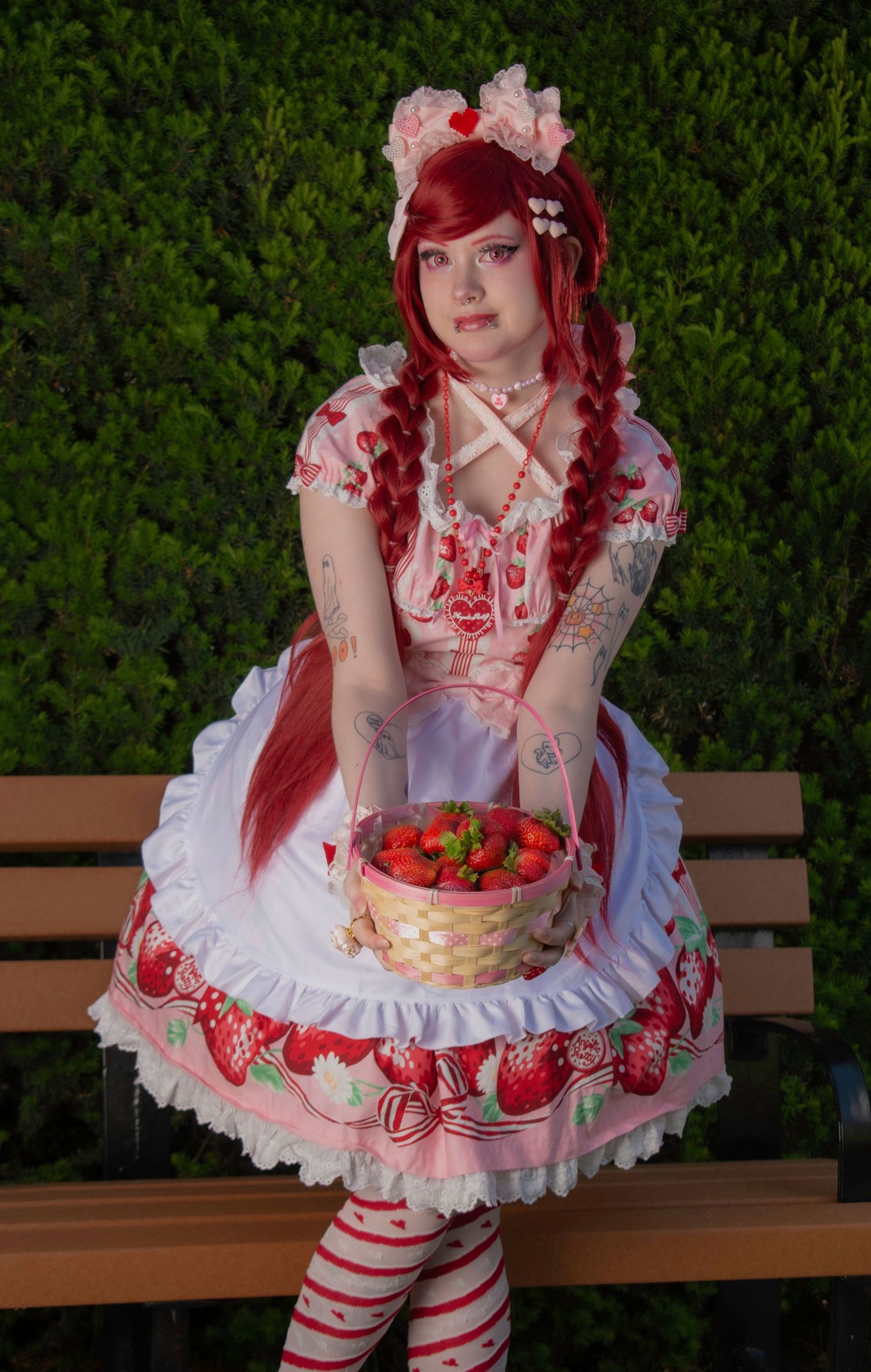 A woman in a strawberry Lolita fashion dress with red braids holding a basket of fresh strawberries on a bench.