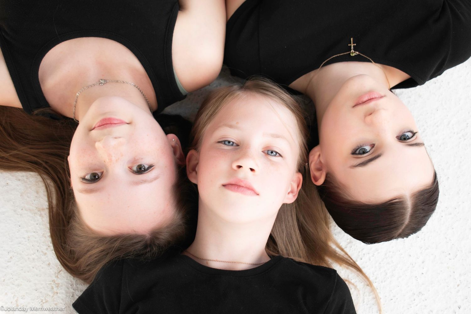 Three children lying on floor with heads together, wearing black shirts, smiling up at camera in a top-down portrait.