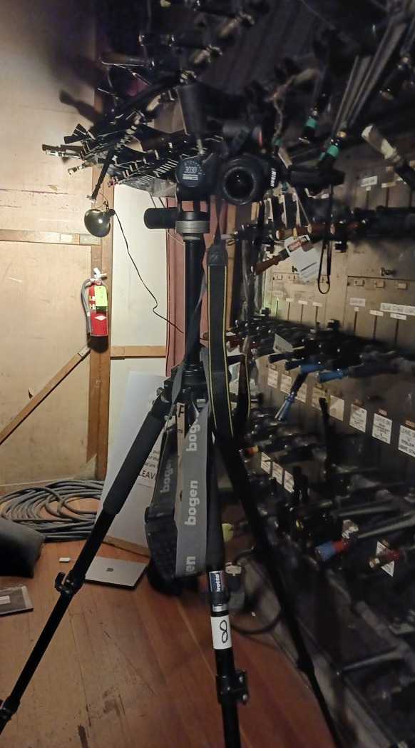 A Bogan camera tripod stands in a storage room surrounded by lighting equipment and cables.