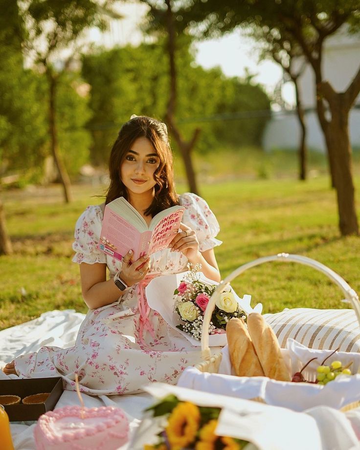A young woman in a floral dress reads a book during a sunny outdoor picnic in a park.