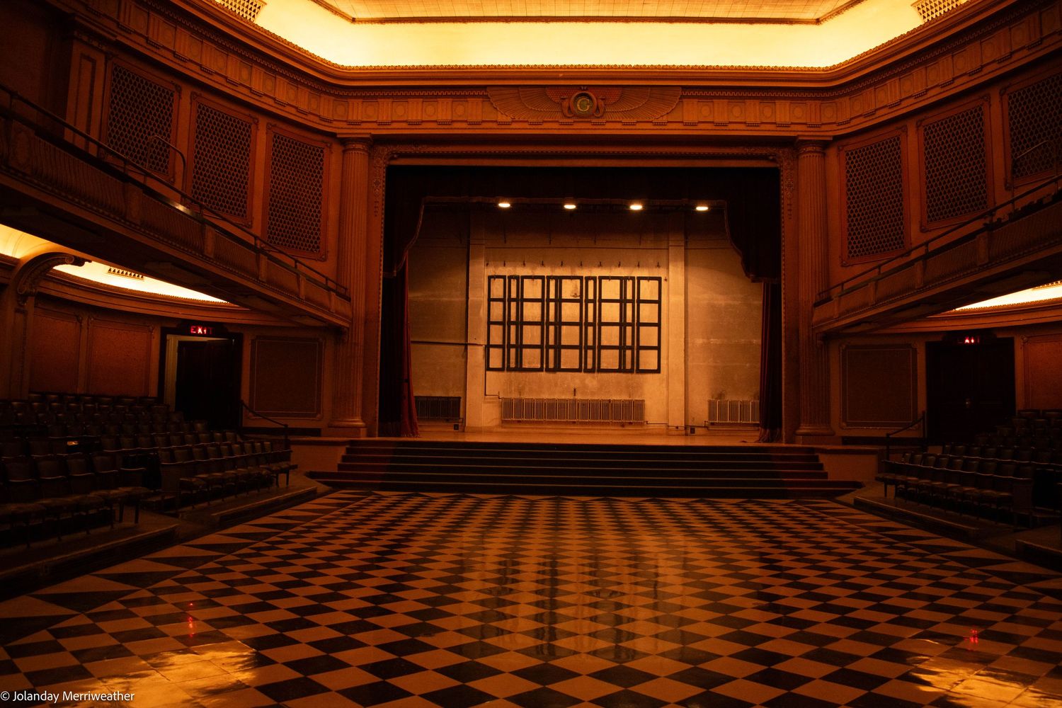 Elegant empty theater with checkered floor, wooden paneled walls, rows of seats, and a lit stage with decorative backdrop.