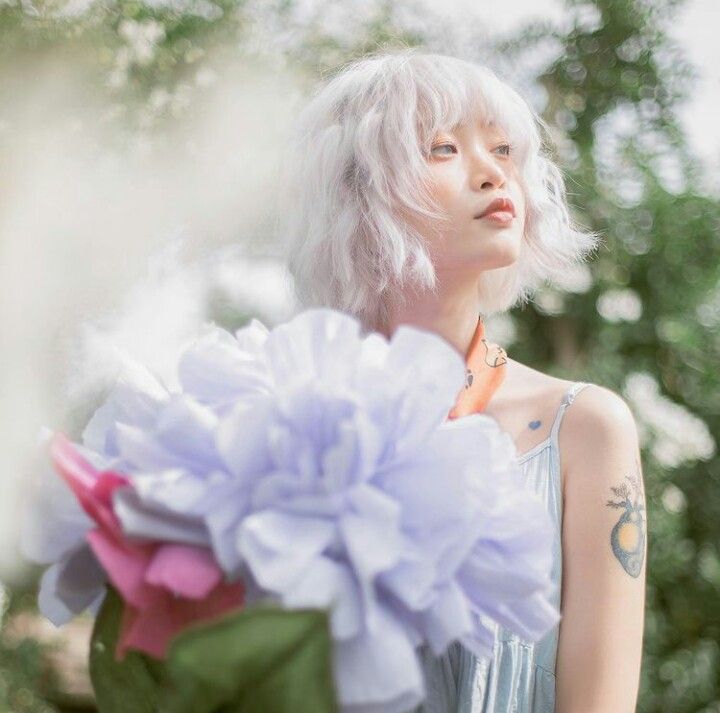 Young woman with silver-white wavy hair holding oversized lavender flowers in a dreamy garden setting.