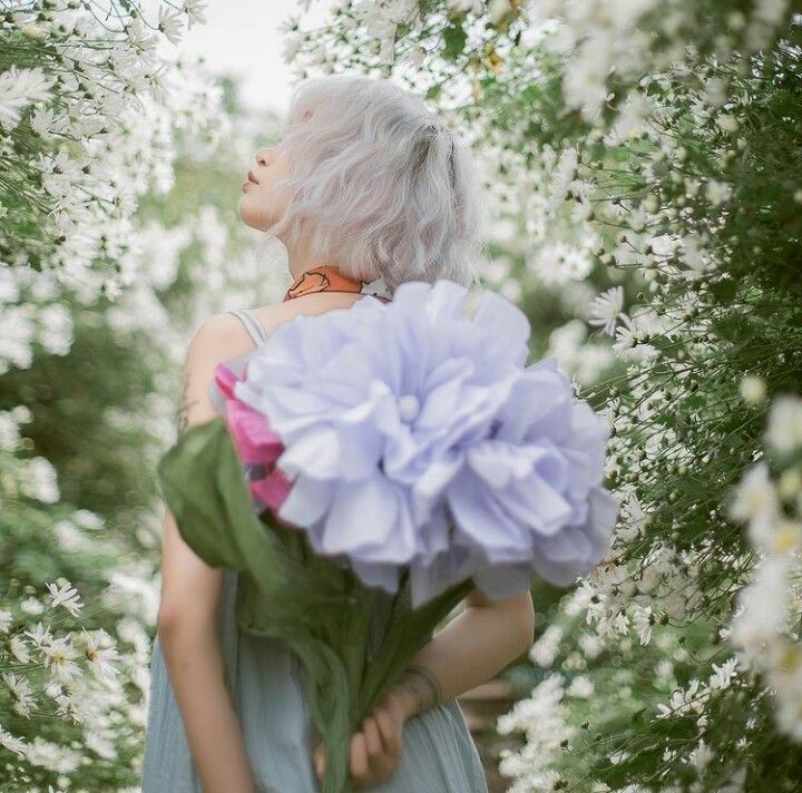 Woman with silver hair holding giant lavender blooms surrounded by white flowering bushes.
