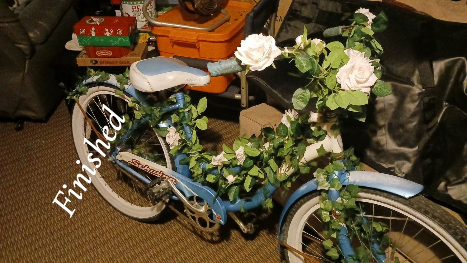 Blue cruiser bicycle decorated with white roses and green ivy garland, labeled Finished, displayed indoors.