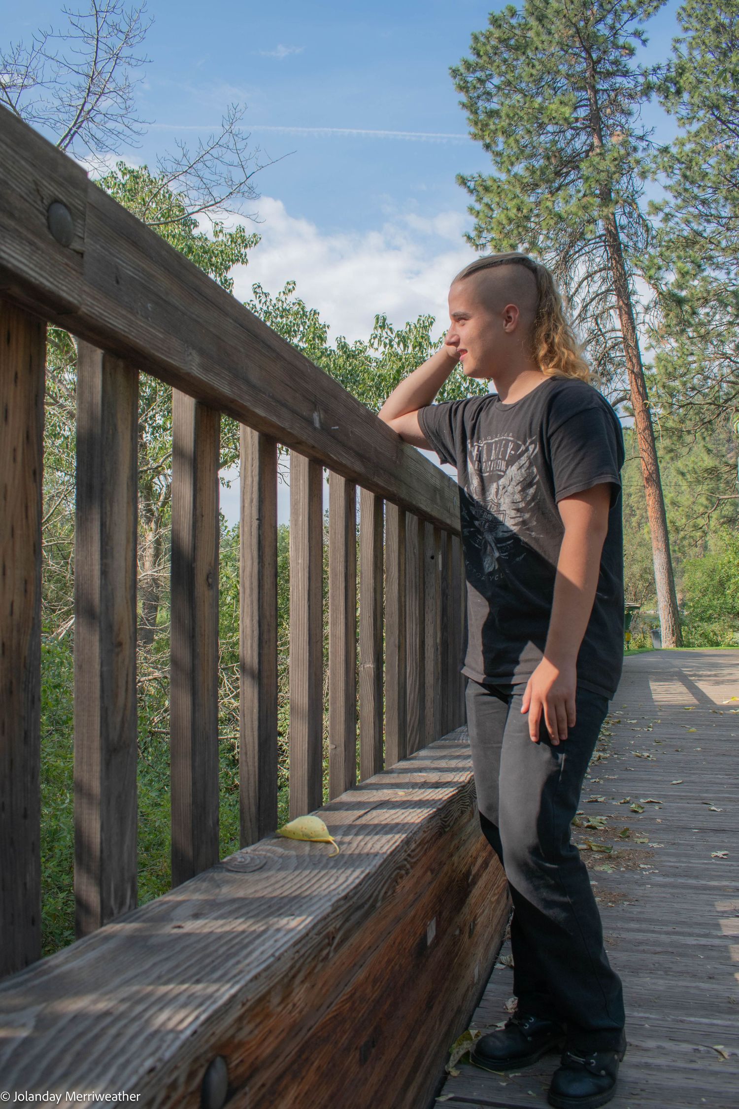 Person with undercut hairstyle leans on a wooden bridge railing overlooking a lush forest trail.