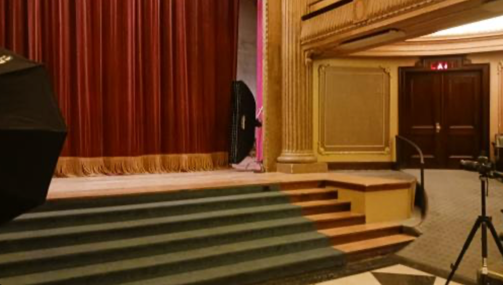 Theater stage with red curtains, wooden steps, and a microphone stand in an elegant auditorium setting.