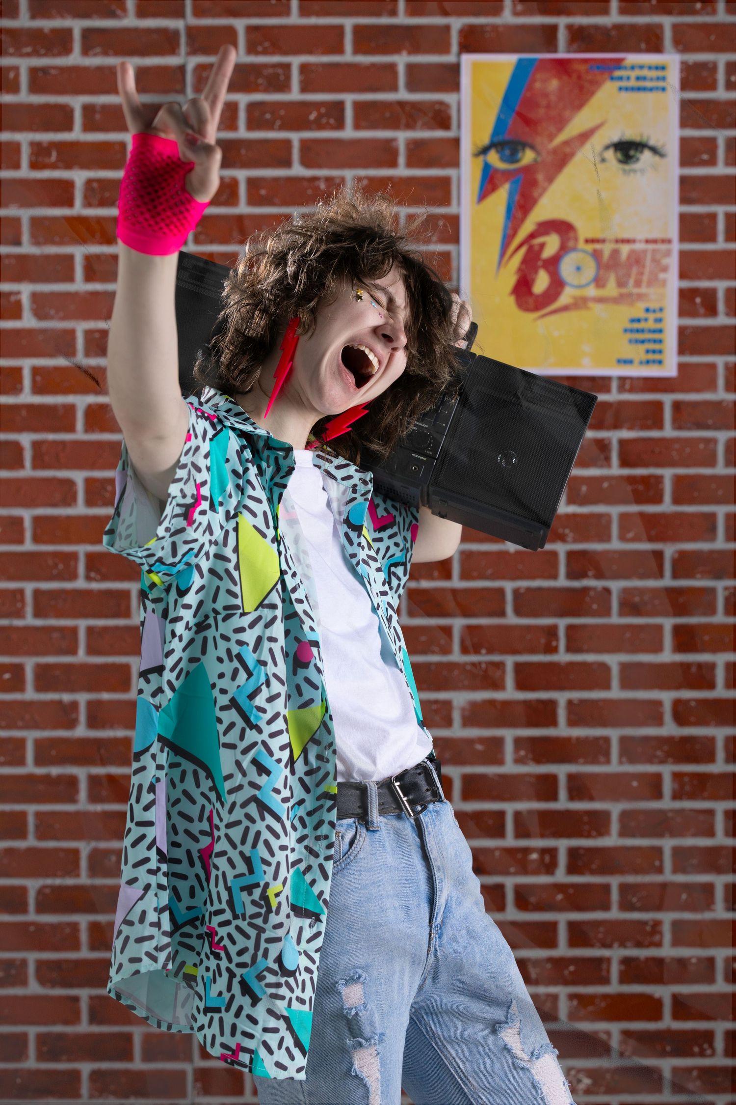 Person in 80s outfit with boombox and rock gesture poses in front of brick wall with David Bowie poster.