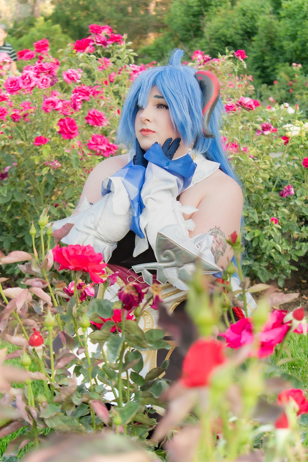 Person in blue wig and anime cosplay costume poses among blooming red roses in an outdoor garden setting.