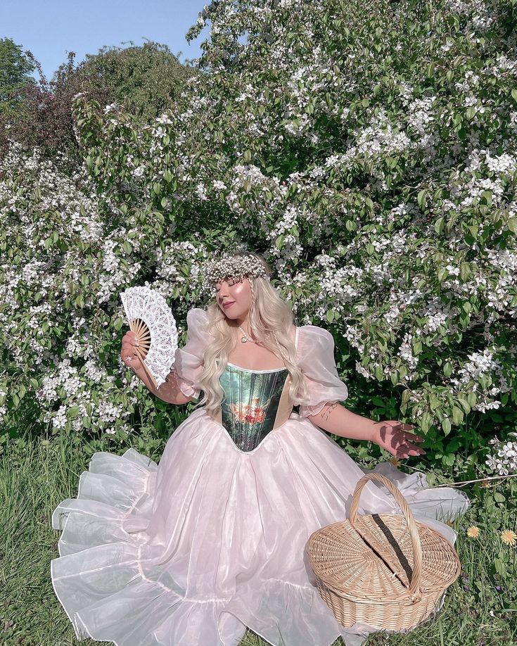 Woman in fairy-tale pink ballgown with floral crown holding lace fan beside blooming white tree.