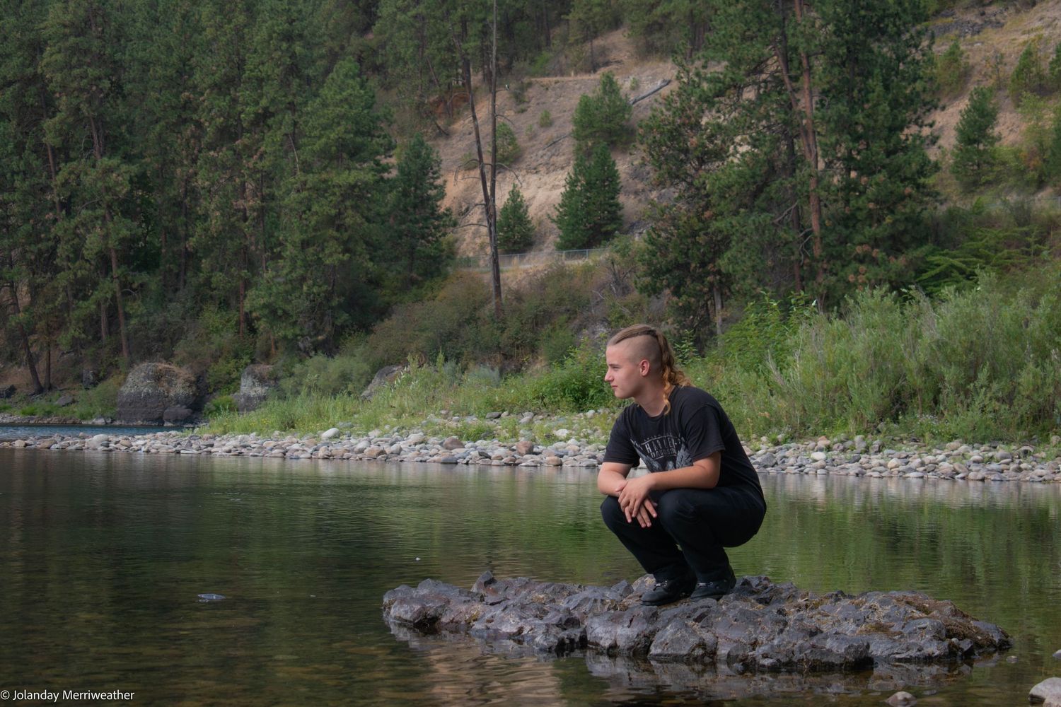 Young person crouches on a rock by a calm river surrounded by pine trees and hillside.