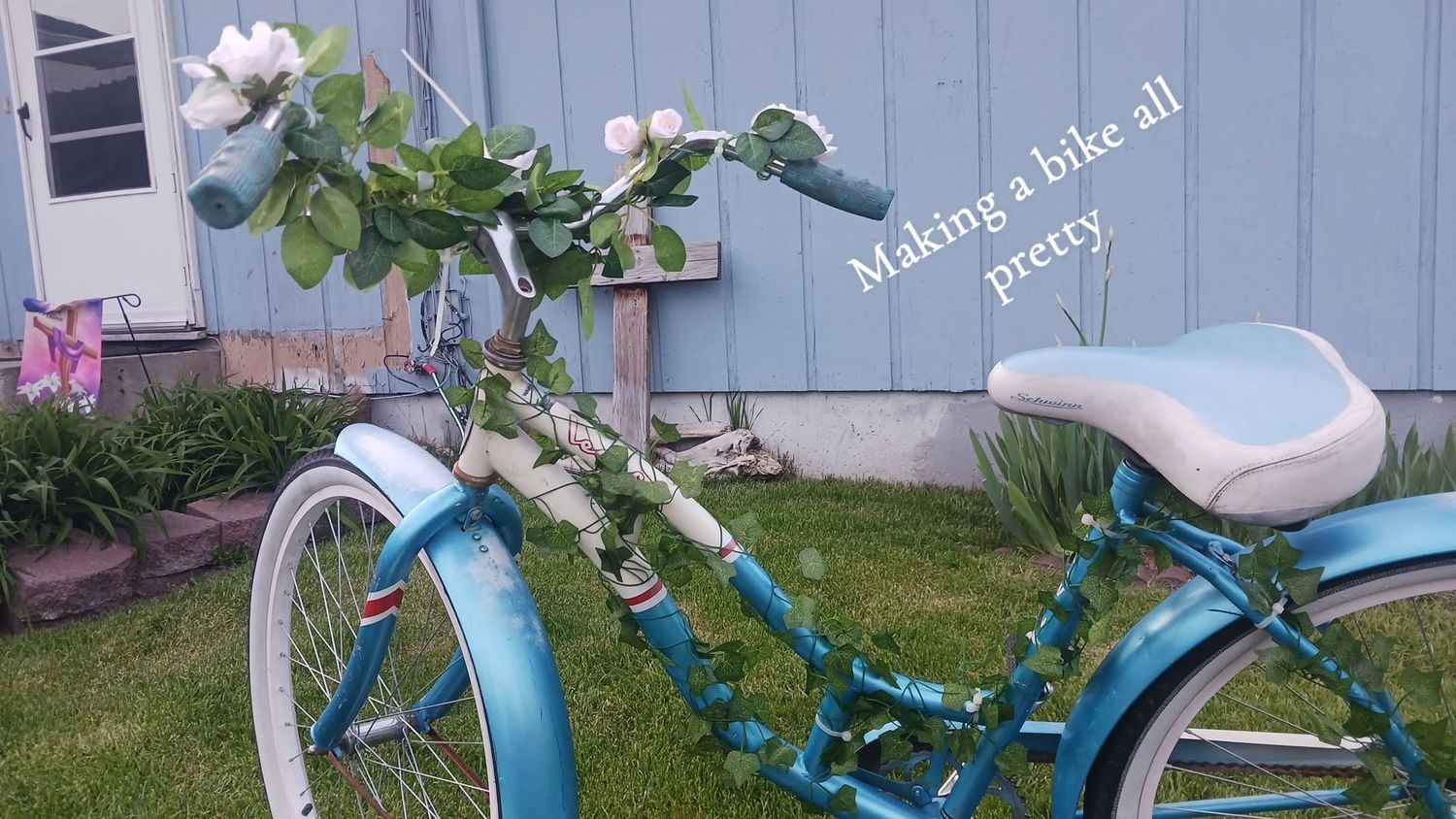 A decorated teal vintage bicycle adorned with green vines and white flowers on the handlebars displayed outdoors.