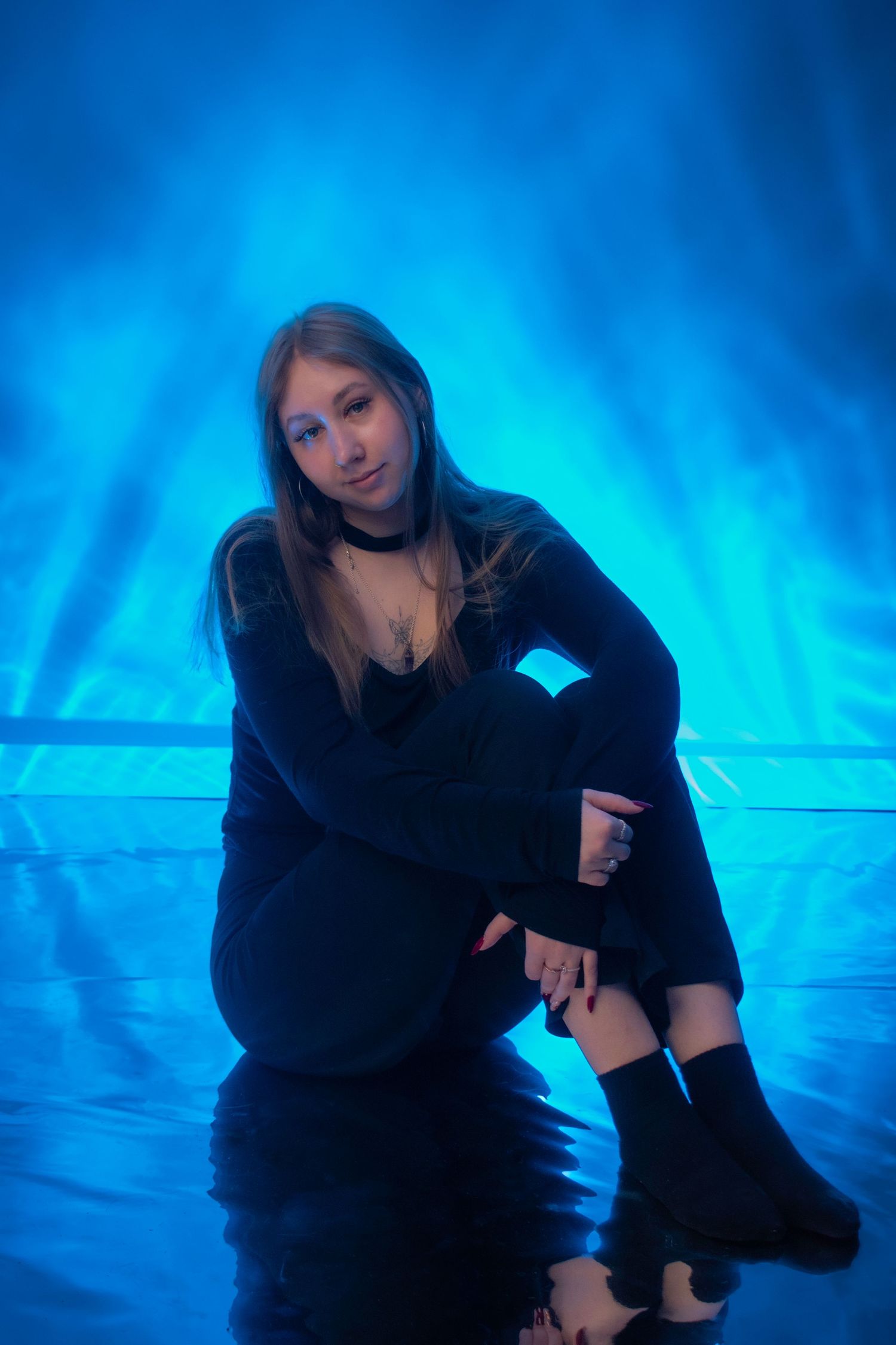 Young woman in black outfit sitting on floor against blue smoky background in dramatic studio photo.