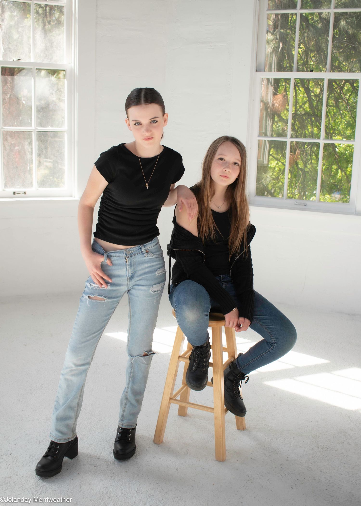 Two young women in black tops and jeans pose together in a bright white studio with large windows.