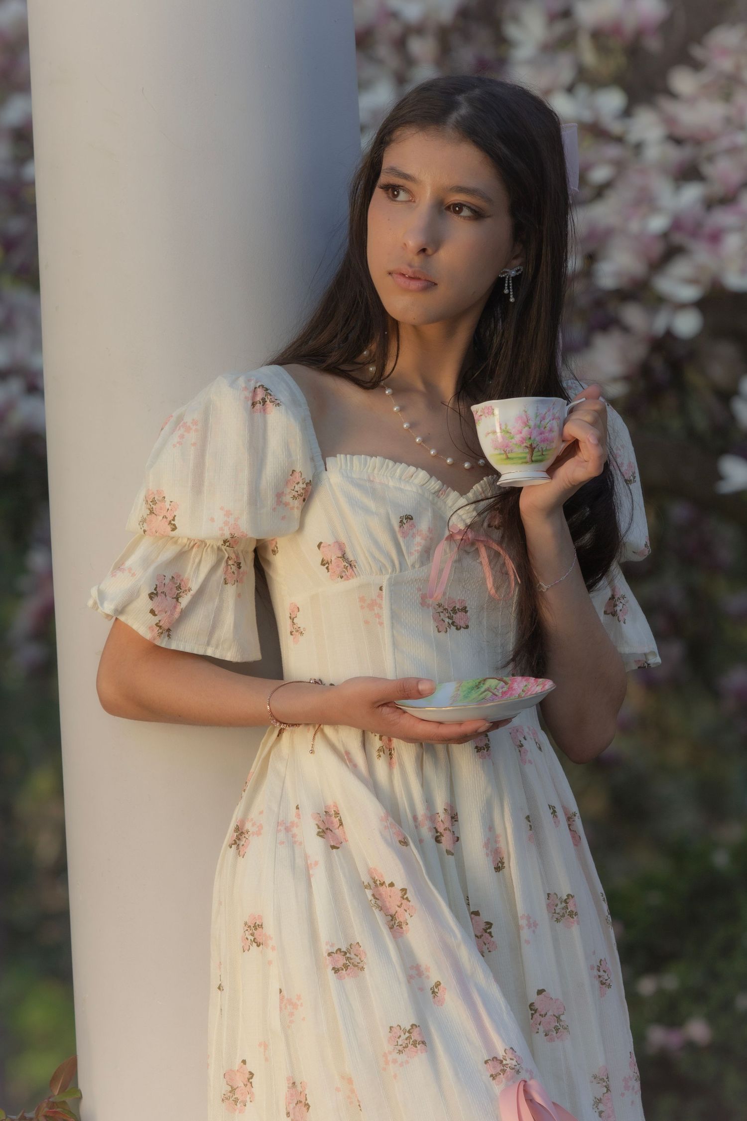 Young woman in a floral dress holding a teacup by a column with blooming magnolia trees in soft natural light.