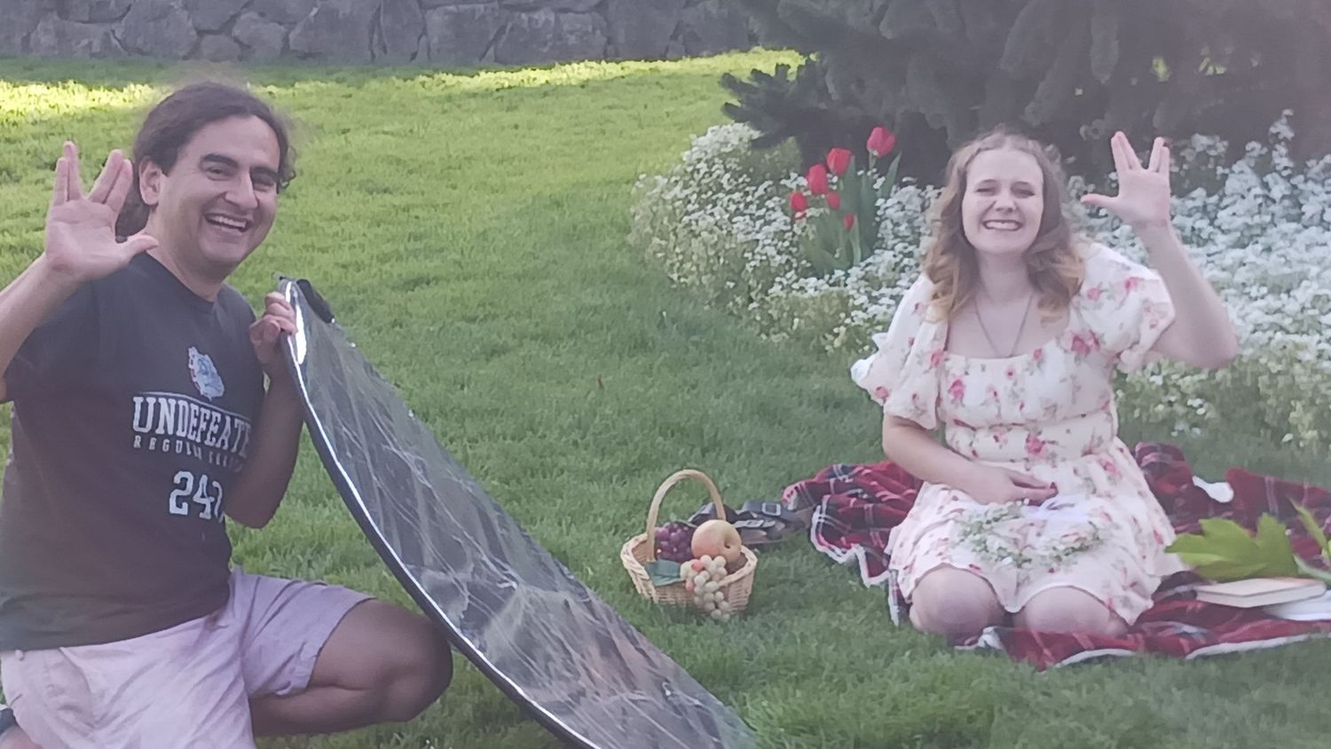 Two people sitting on a picnic blanket in a garden, smiling and making peace signs with their hands.
