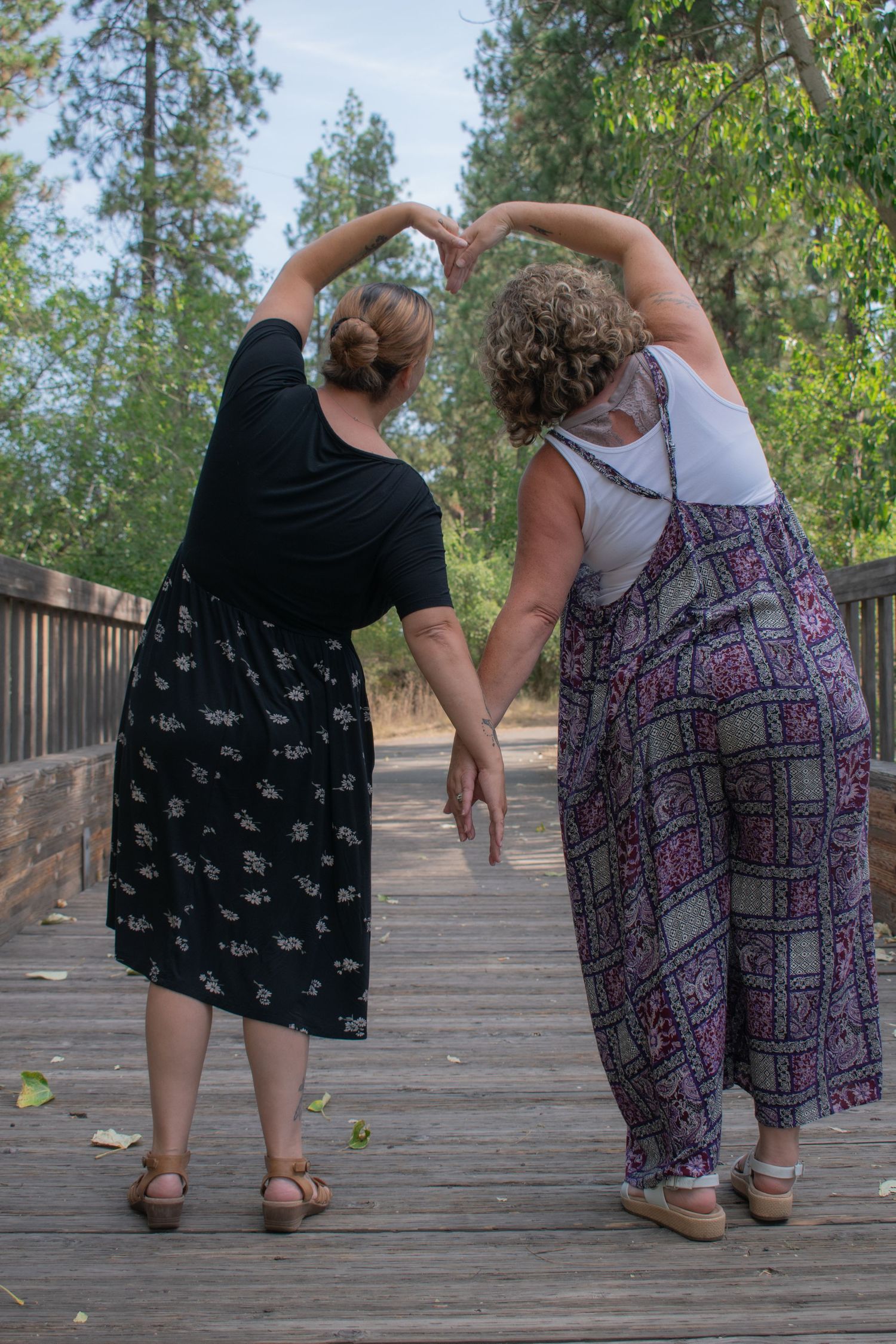 Two women forming a heart shape with their arms on a wooden bridge path.