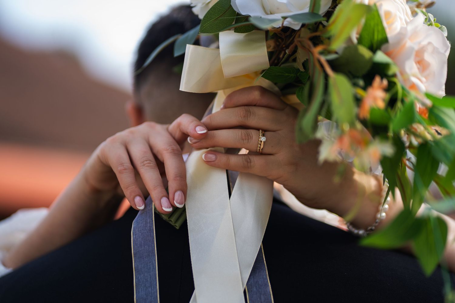 Close-up of hands holding a wedding bouquet wrapped in white ribbon, showing an engagement ring and manicured nails.