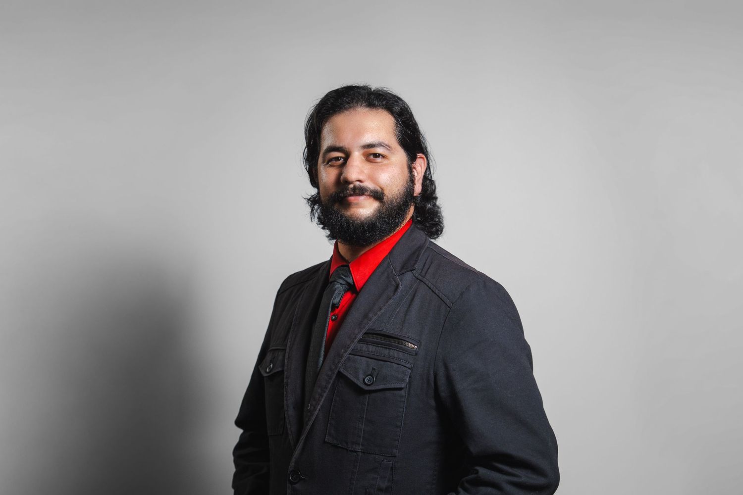 Side-profile professional headshot of man in black suit with red shirt on gray background