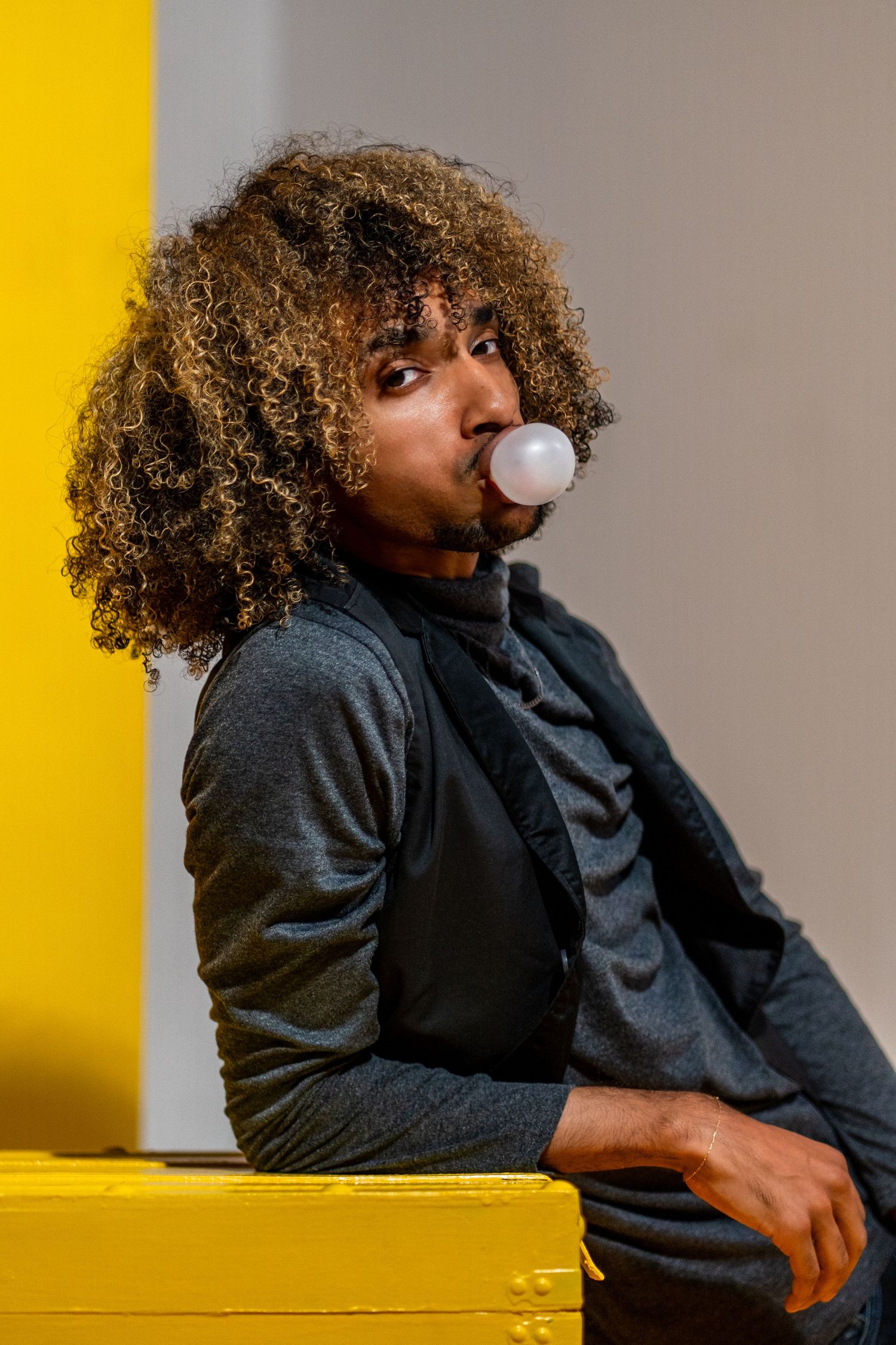 Playful  bold fashion portrait in Wynwood, Miami, FL – stylish male model with curly hair blows a bubblegum bubble against a bright yellow