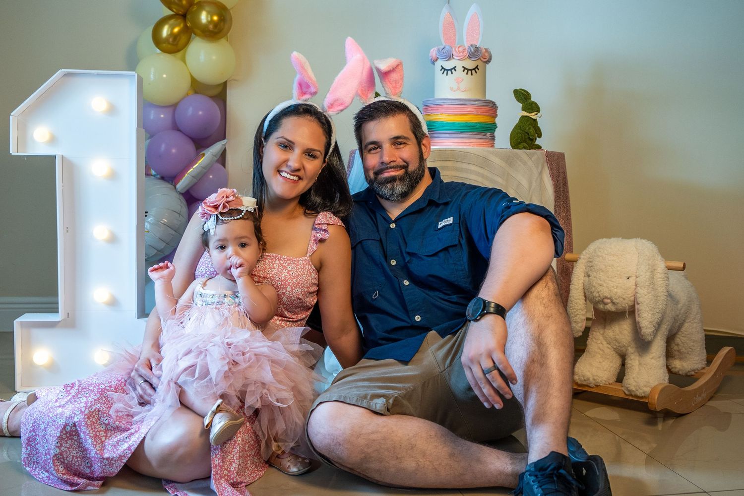 Parents sitting with their baby girl beside a lit number one and balloon display during an indoor first birthday session.