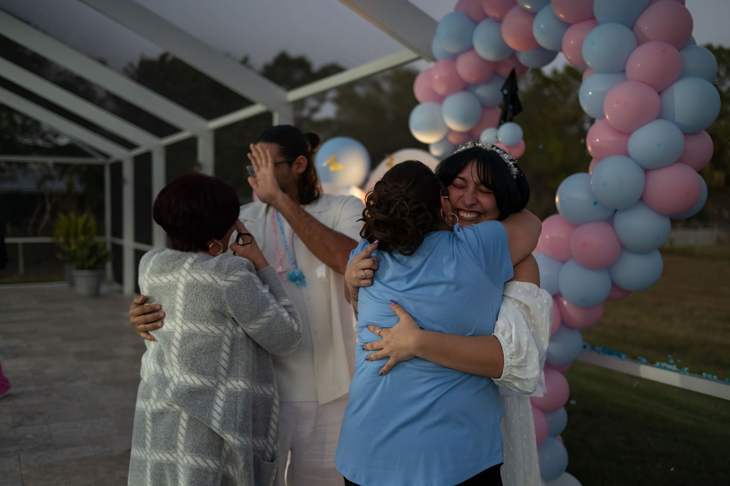 Family hugging and celebrating a gender reveal under a balloon arch at an outdoor South Florida event.