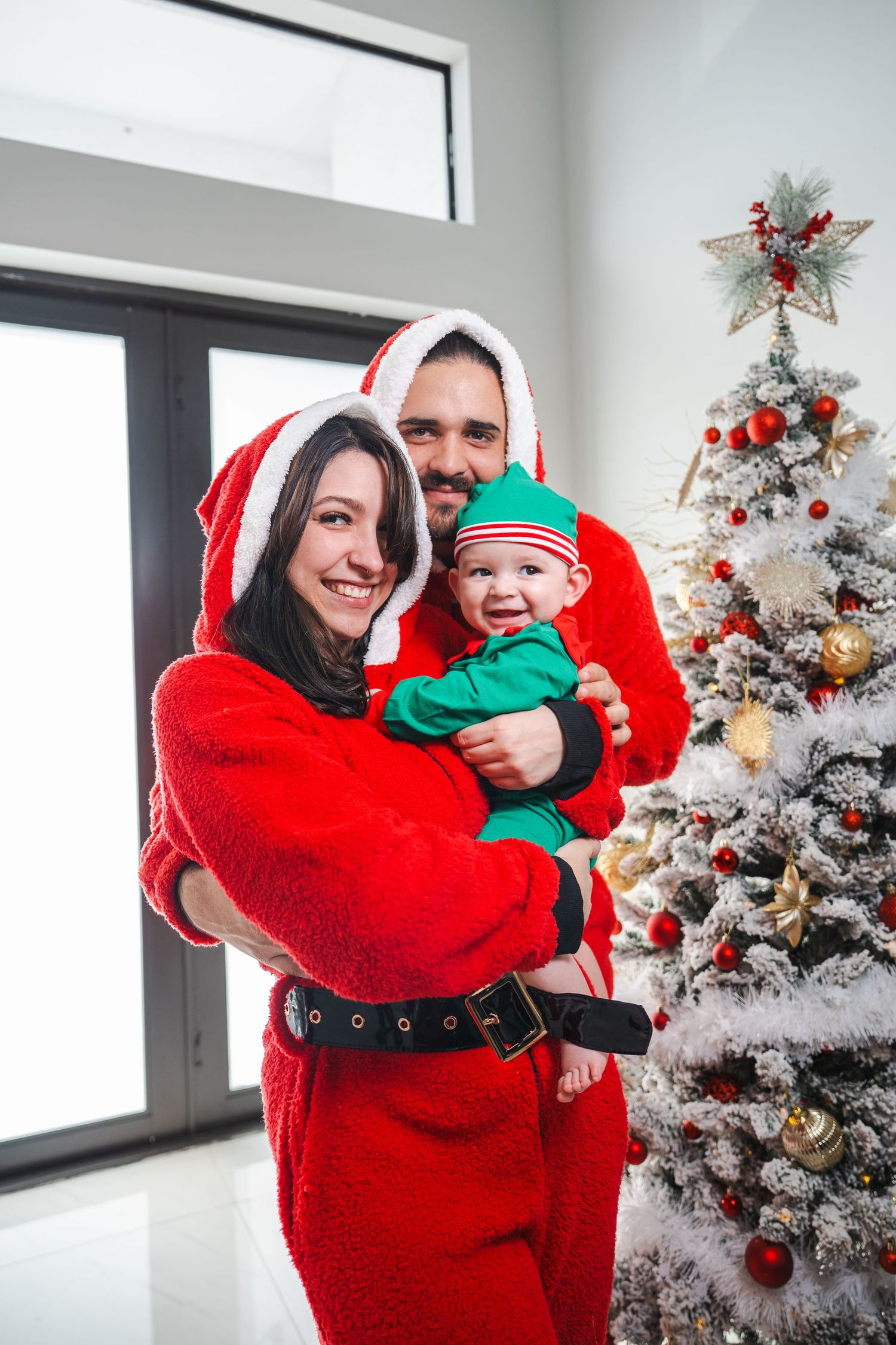 Family Christmas portrait with parents and baby by a decorated tree, photographed indoors in South Florida.