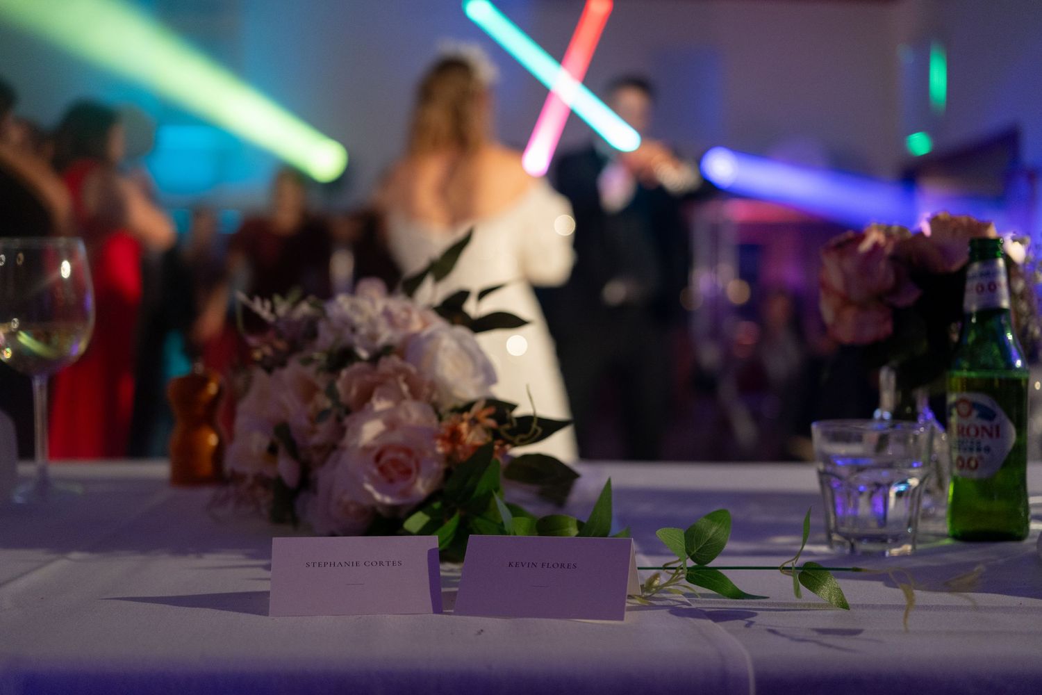 Documentary-style wedding reception photo capturing a couple dancing under colorful lights, with floral details in the foreground.