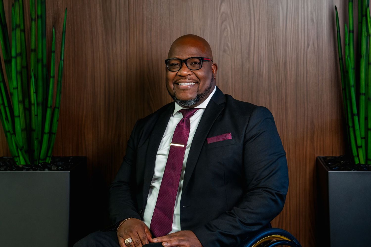 Professional wearing a dark suit and burgundy tie sits at desk with green plants and wood paneling in background.
