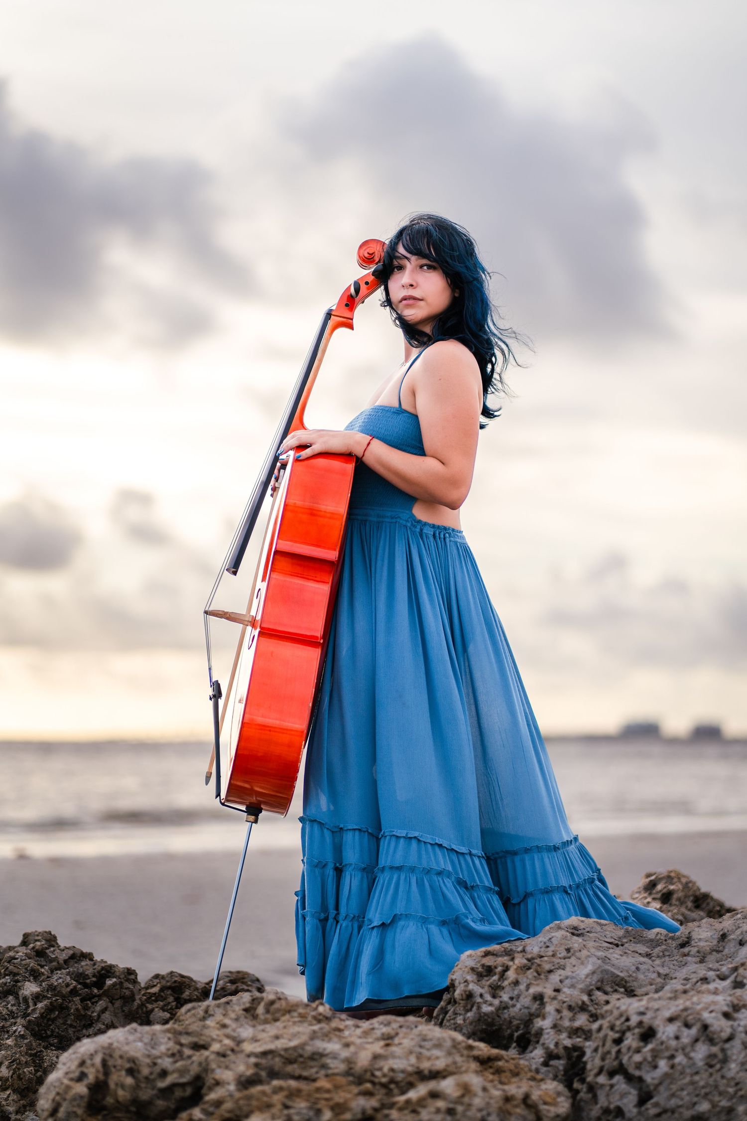 Cellist in blue dress posing on rocks during creative branding session in South Florida