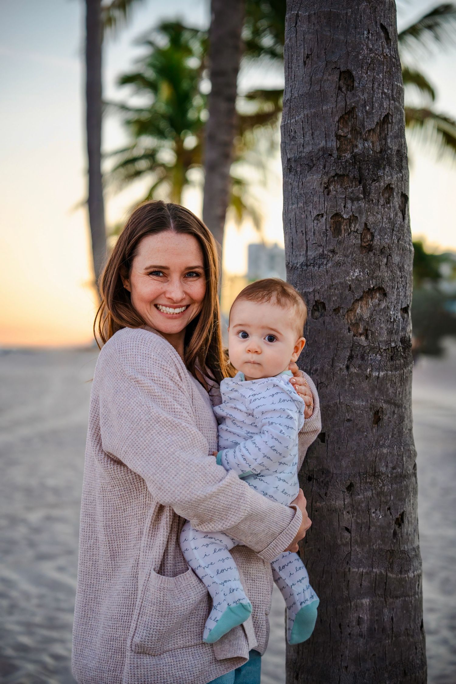 Mother holding her baby beside a palm tree at sunset during a relaxed South Florida beach family session.