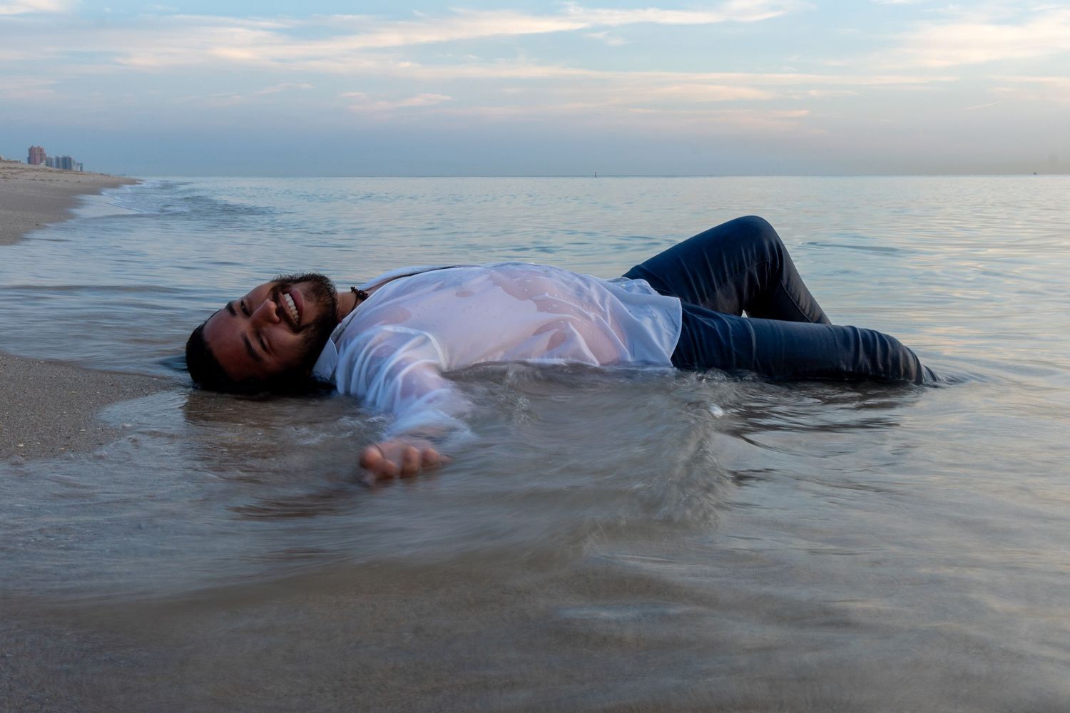 Artistic beach session – man in a soaked white shirt lies in the sand as waves wash over him, capturing a cinematic mood.