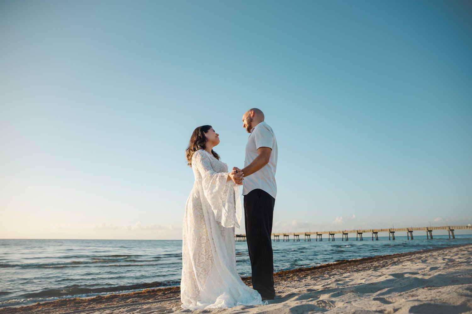 Couple standing together on beach with pier in background during maternity session