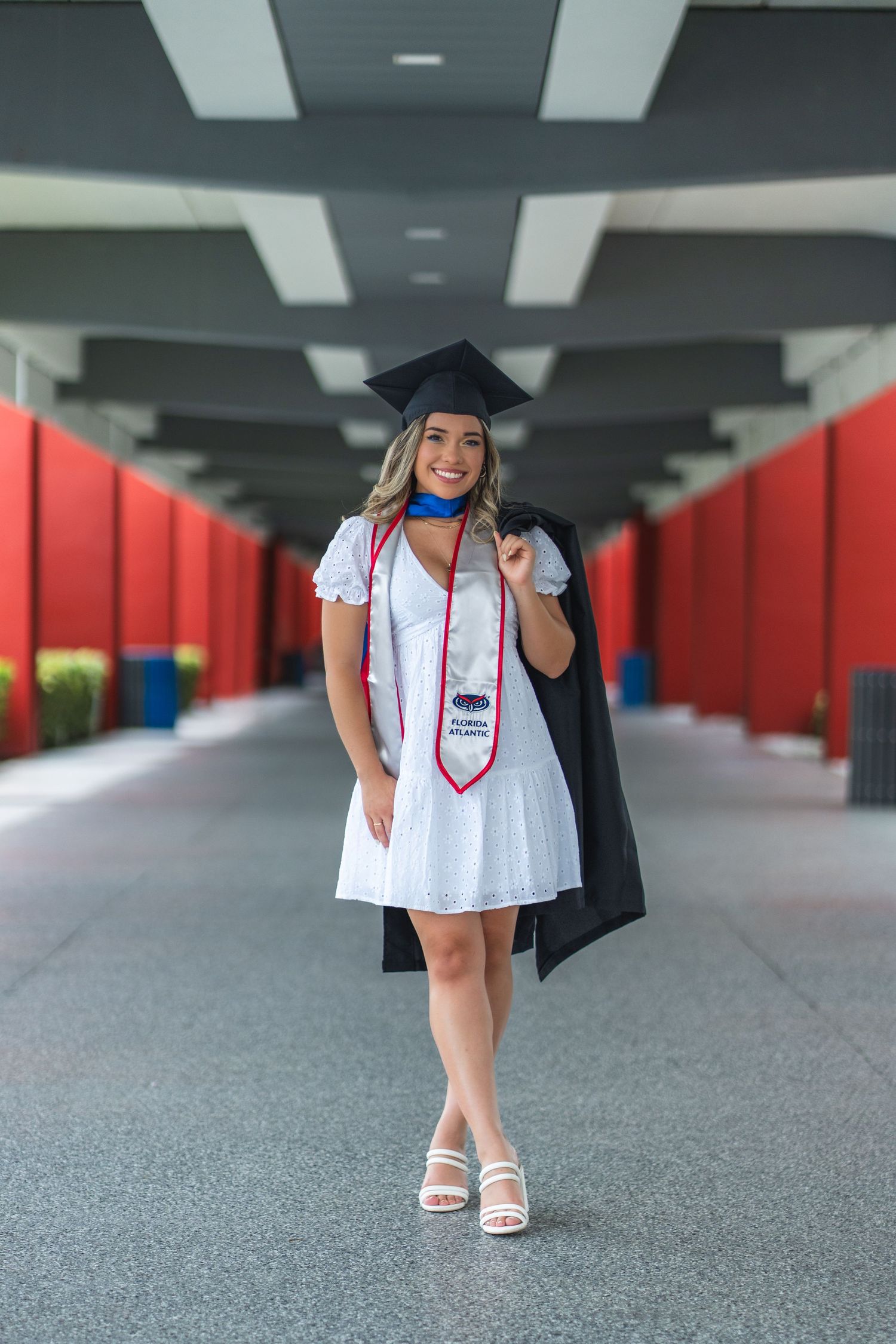 Proud FAU graduate posing in her white dress and black graduation gown under a red and black structure at Florida Atlantic University.