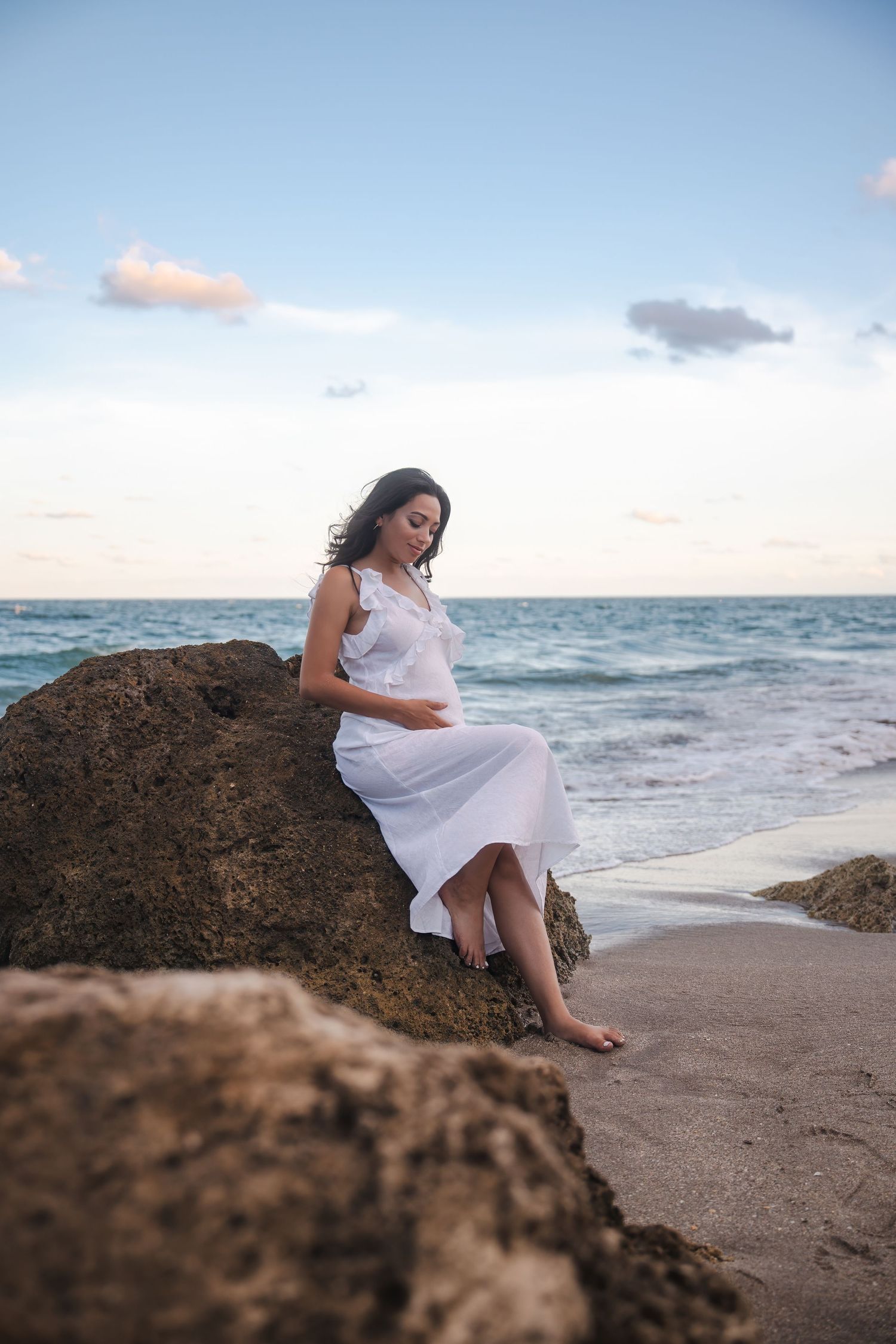 Beach pregnancy announcement portrait of an expecting mother seated on coastal rocks at sunset in Deerfield Beach, Florida